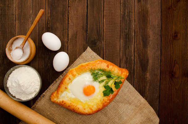 Khachapuri with eggs on sackcloth, salt, flour, eggs and parsley on the wooden table, space for text - Photo, Image