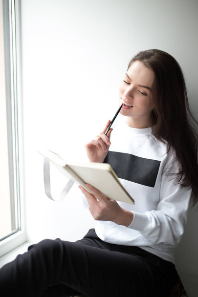 Beautiful young woman sitting with notebook on windowsill - Foto, afbeelding