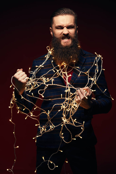 Porträt eines modischen Mannes mit Bart und Weihnachtsgirlanden im Atelier  - Foto, Bild