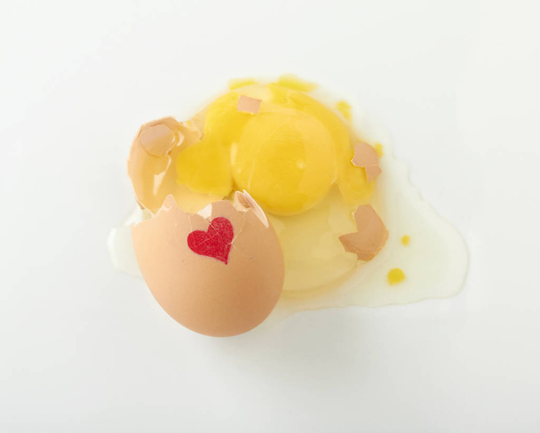 Beyaz bir yüzey üzerinde kırmızı bir kalp ile kırık çiğ yumurta - Fotoğraf, Görsel