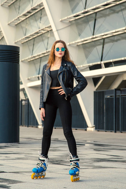 Girl on rollerblades standing in building background. Young fit women girl in blue sunglasses, jeans and jacket on roller skates riding outdoors after rain. - Foto, afbeelding