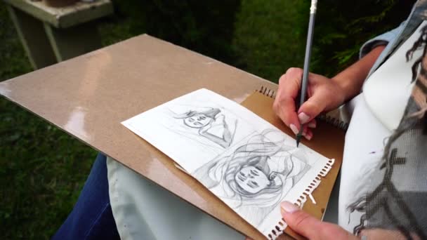 Женщина держит серый карандаш в руке, делает эскизы и контур на листе бумаги и сидит в парке на открытом воздухе
. - Кадры, видео