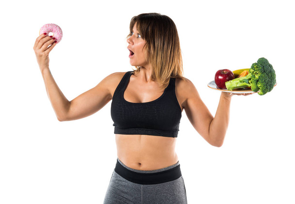 Спортивная женщина держит фрукты и овощи в одной руке и пончик в другой руке
 - Фото, изображение