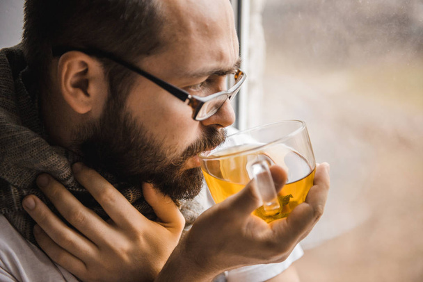 άρρωστος άνθρωπος με ένα μαντήλι γύρω από το λαιμό του πίνοντας τσάι στο σπίτι κάθεται στο παράθυρο - Φωτογραφία, εικόνα