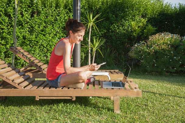 vrouw met telefoon en computer op lounge houten strandstoel - Foto, afbeelding