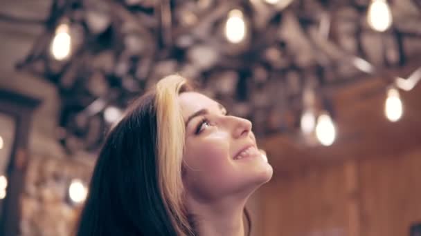 Young brunette woman looks up at the ceiling decorated with lamps and turns to camera - Séquence, vidéo