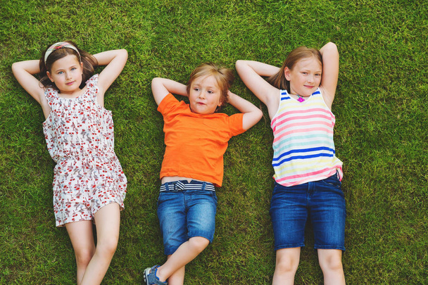 Щасливі діти розважаються на відкритому повітрі. Діти грають у літньому парку. Маленький хлопчик і дві дівчинки лежать на зеленій свіжій траві
 - Фото, зображення