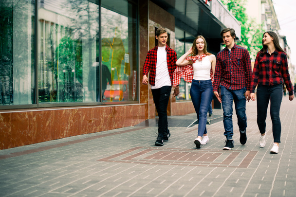 Four happy trendy teenage friends walking in the city, talking each other and smiling. Lifestyle, friendship and urban life concepts. - Фото, изображение