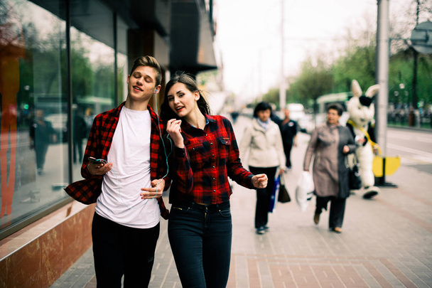 Two happy trendy teenage friends walking and dancing in the city, listening to the music with headphones, talking each other and smiling. Lifestyle, friendship and urban life concepts. - Photo, Image