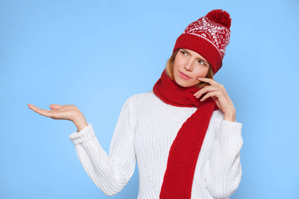 Young beautiful woman thinking looking to the side with showing open hand palm at blank copy space, Christmas girl wearing knitted warm hat and scarf, isolated over blue background - Foto, afbeelding