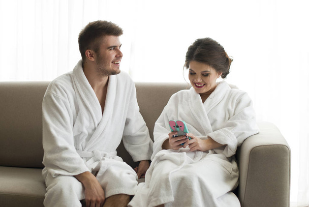 Atractiva pareja joven está mirando el teléfono inteligente en el sofá en el albornoz
 - Foto, imagen