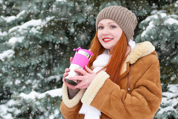 hermosa mujer bebiendo una bebida caliente y mantener caliente en invierno al aire libre, abetos nevados en el bosque, pelo largo y rojo, con un abrigo de piel de oveja
 - Foto, Imagen