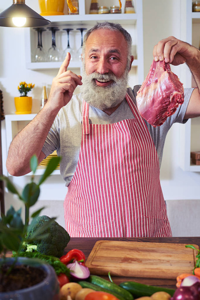 Homem com um pedaço de carne crua a gesticular o polegar na cozinha
 - Foto, Imagem