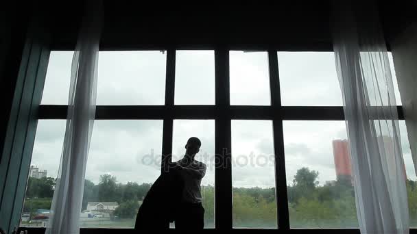 Ο άνθρωπος φορέματα βάζοντας στο κοστούμι silhouetted φωτιζόμενη στο μεγάλο παράθυρο. Προετοιμασία για το επίσημο βραδινό ή γαμήλια τελετή - Πλάνα, βίντεο