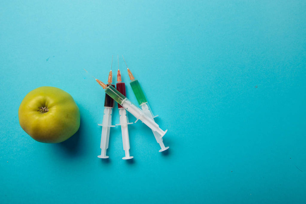 Chemical additives in food or genetically modified fruit concept. Green apple with syringes of chemicals. Isolated on blue background. - Photo, Image