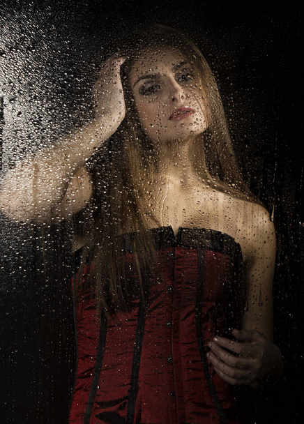 Portrait of a sensual young sexy woman wearing corset, touching her hair and posing behind transparent glass covered by water drops. - Foto, Bild