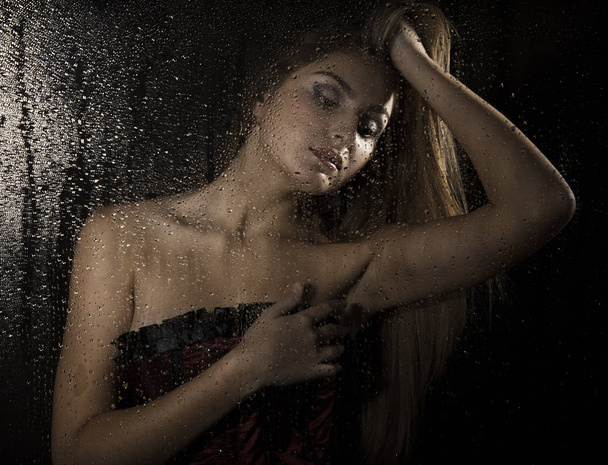 Portrait of a sensual young sexy woman wearing corset, touching her hair and posing behind transparent glass covered by water drops. - Fotó, kép