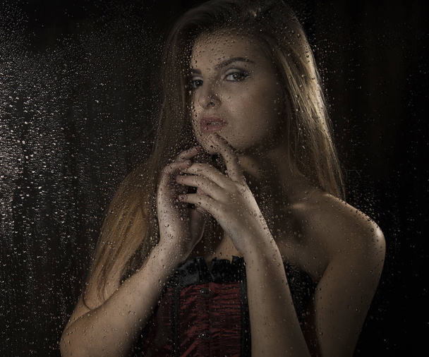Portrait of a sensual young sexy woman wearing corset, touching her hair and posing behind transparent glass covered by water drops. - Photo, image