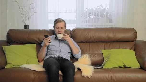Young man drinking tea, listening to music and singing - Záběry, video