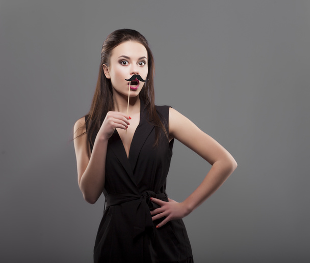 Girl holding funny paper mustache, isolated, gray background. Shoked young fashion woman. - Фото, изображение