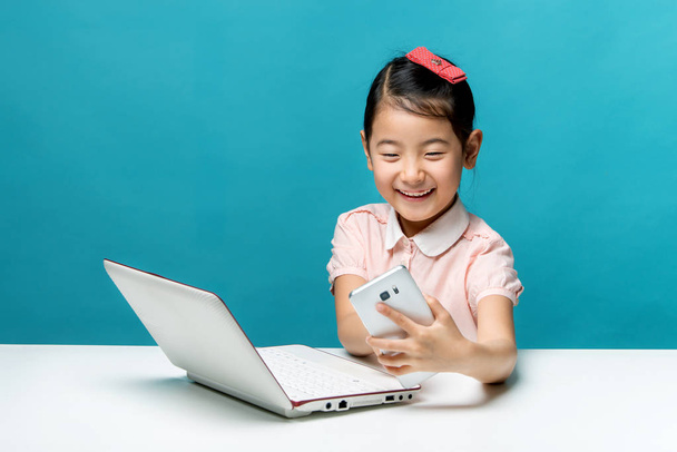 Cute asia little girl is sitting at table with her white laptop and a smartphones, isolated over blue - Фото, изображение