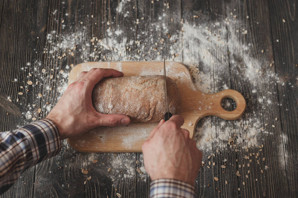 Baking and cooking concept background. Hands of baker closeup cutting bread loaf with knife on rustic wooden table sprinkled  flour. - Photo, Image