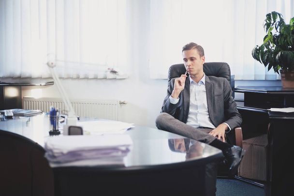 Αρσενικά ενήλικα άνθρωπος σε ένα κοστούμι που κάθεται σε μια καρέκλα πίσω από το γυαλιστερό τραπέζι σε ένα γραφείο, να σκέφτεται - Φωτογραφία, εικόνα