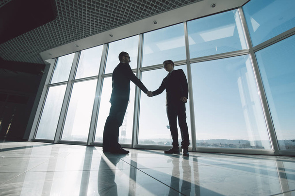 Dois jovens empresários estão apertando as mãos um com o outro em pé contra as janelas panorâmicas
. - Foto, Imagem