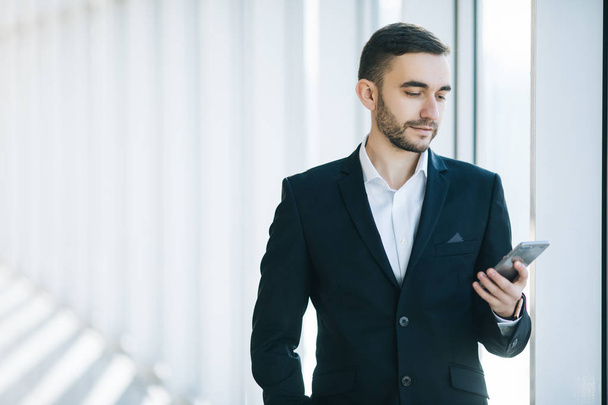 Bello uomo d'affari in giacca e cravatta guarda smartphone in ufficio
 - Foto, immagini