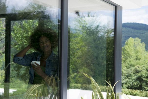 African American vrouw drinken koffie kijkt uit het raam - Foto, afbeelding