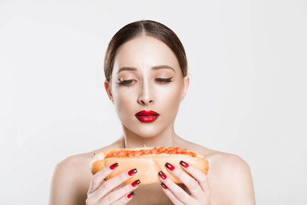 Not healthy. Young beautiful woman wants to eat hotdog craving but she stops because it is unhealthy food. Natural red lips black eyeliner girl, false eyelashes makeup isolated over white background. - Foto, immagini