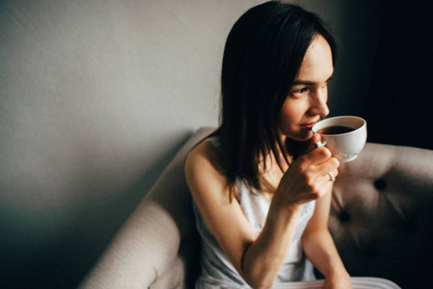 Nainen pyjamassa kahvikupin kanssa istuu nojatuolilla kotona
  - Valokuva, kuva