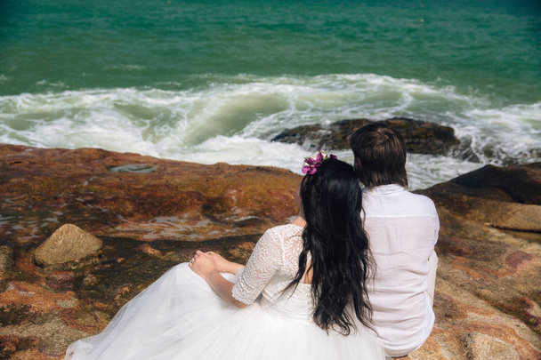 наречений в білому костюмі і наречена в сукні тримає один одного руками, стоячи на тлі моря. красивий пейзаж з великими каменями. концепт день весілля
 - Фото, зображення