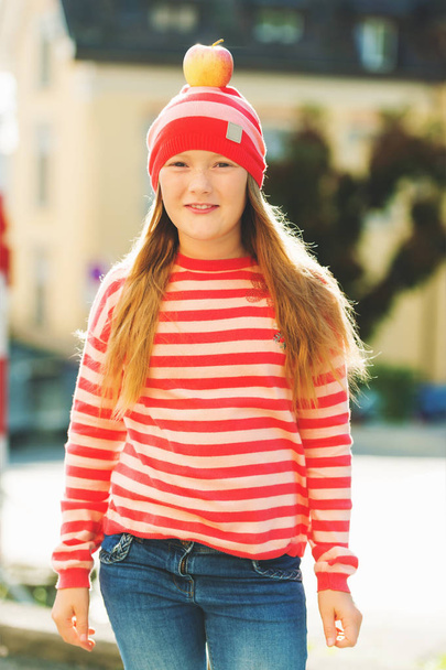 Retrato ao ar livre da jovem menina de 9-10 anos, vestindo chapéu listra vermelha e pulôver, maçã na cabeça
 - Foto, Imagem