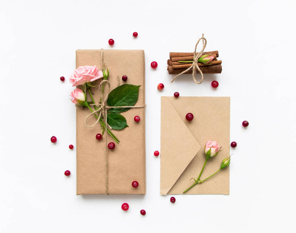 Gift box and envelope in eco paper on white background. Presents decorated with roses and berries. Holiday concept, top view, flat lay - Photo, Image