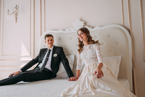 roztomilý svatební pár v interiéru ateliéru klasické bílé pózuje na posteli zdobené. Hej políbit a obejmout, drželi se za ruce na sebe dívali - Fotografie, Obrázek