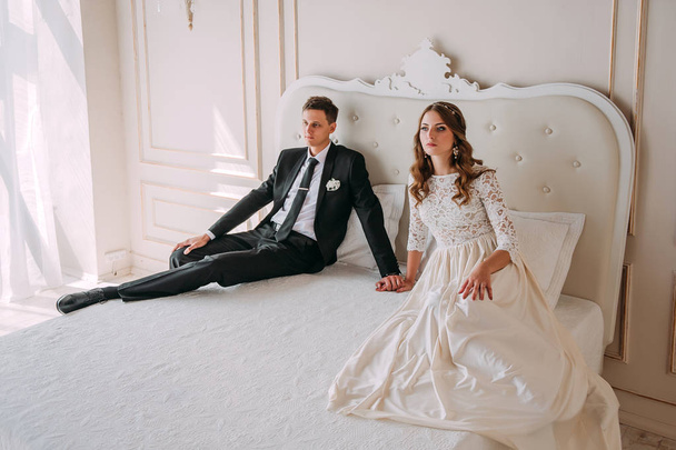 schattig bruidspaar in het interieur van een klassieke witte studio poseren op een bed ingericht. Hey kussen en omhelzen elkaar, hand in hand kijken naar elkaar - Foto, afbeelding