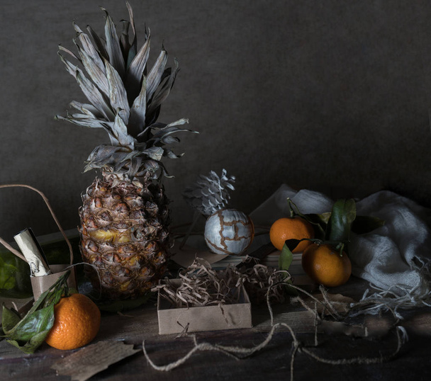 still life, vintage. pineapple, tangerines, boxes, scarf, decor on a wooden table, dark background. space for text - Foto, Bild