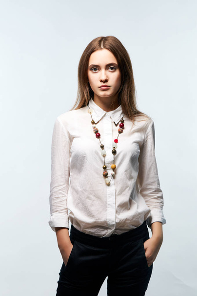 Mujer joven con estilo en pantalones negros, camisa blanca y decoraciones masivas en el cuello. Retrato de estudio de una joven empresaria
. - Foto, imagen