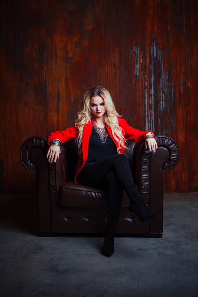 Young and attractive blond woman in red jacket sits in leather armchair, background grunge rusty wall - Foto, imagen