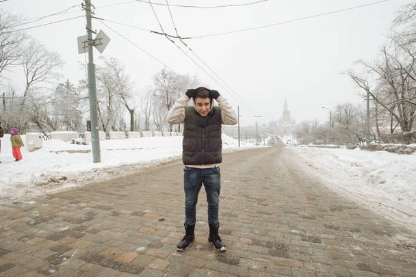 Outdoor winter portrait for young handsome man. Beautiful teenager in his jacket and vest posing on a city street, background of fir trees. - Zdjęcie, obraz