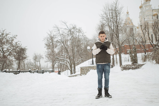 Outdoor winter portrait for young handsome man. Beautiful teenager in his jacket and vest posing on a city street, background of fir trees. - Fotó, kép
