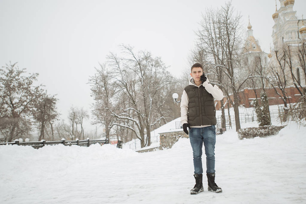 Outdoor winter portrait for young handsome man. Beautiful teenager in his jacket and vest posing on a city street, background of fir trees. - Photo, Image