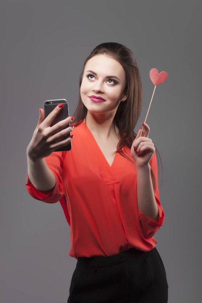 Girl in red shirt, gray background. Joyful young fashion brunette woman doing selfie with smartphone, holds paper heart. Positive emotion feeling, smiling. - Foto, Imagem
