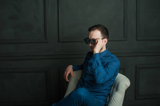 studio portret van een jonge hipster man op een donkere muur achtergrond in een blauw shirt en spijkerbroek.  - Foto, afbeelding