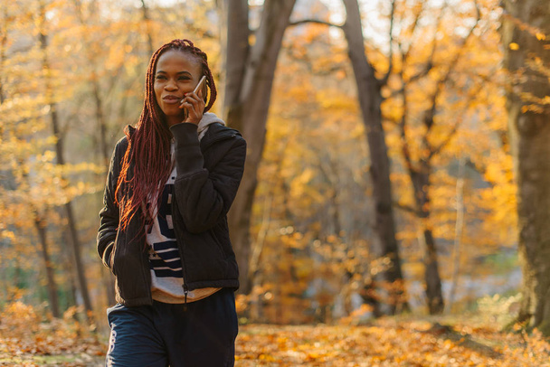 Cute african girl walking in autumn park and talking by phone. Woman have a rest on fresh air. - Fotó, kép