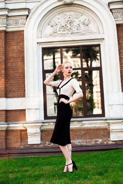 Happy Fashion blonde european elegant Woman with red lips and white skin Standing at the Old Red Brick building on green grass. Glamour Girl Outdoors. Beautiful Female in formal black and white dress. - Photo, Image