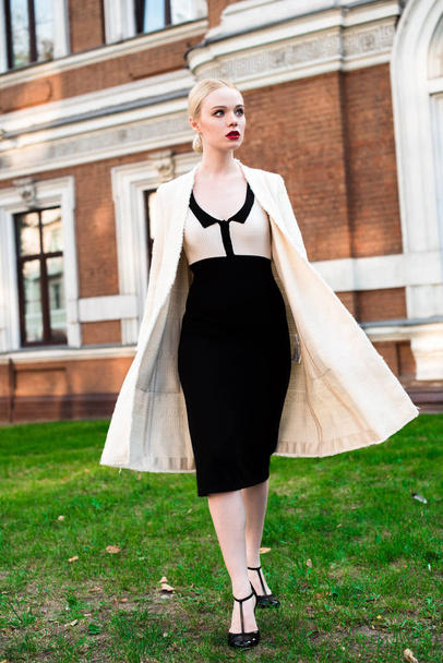 Happy Fashion blonde european elegant Woman with red lips and white skin Standing at the Old Red Brick building on green grass. Glamour Girl Outdoors. Beautiful Female in formal black and white dress. - Photo, image