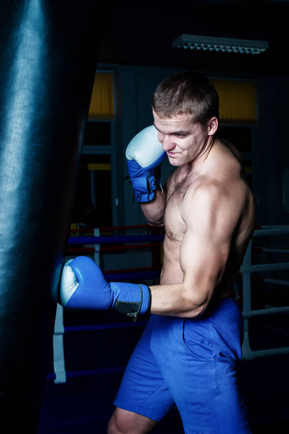 Handsome man in blue boxing gloves training on a punching bag in the gym. Male boxer doing workout. - Foto, afbeelding
