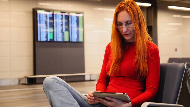 Young attractive woman with red hair and glasses use gadget in airport departure lounge, close up - 写真・画像